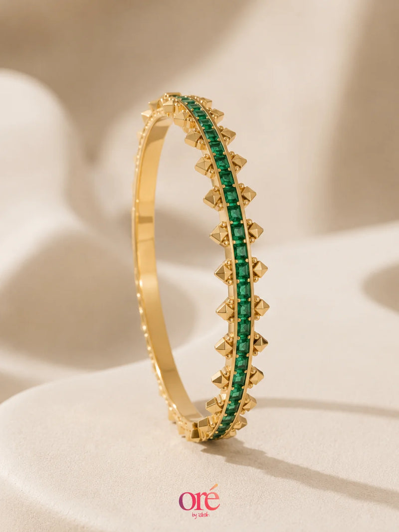 A gold bangle bracelet featuring a central row of emerald-cut green stones, standing upright on a soft beige fabric surface. The brand logo 'ore by ore' is visible at the bottom.