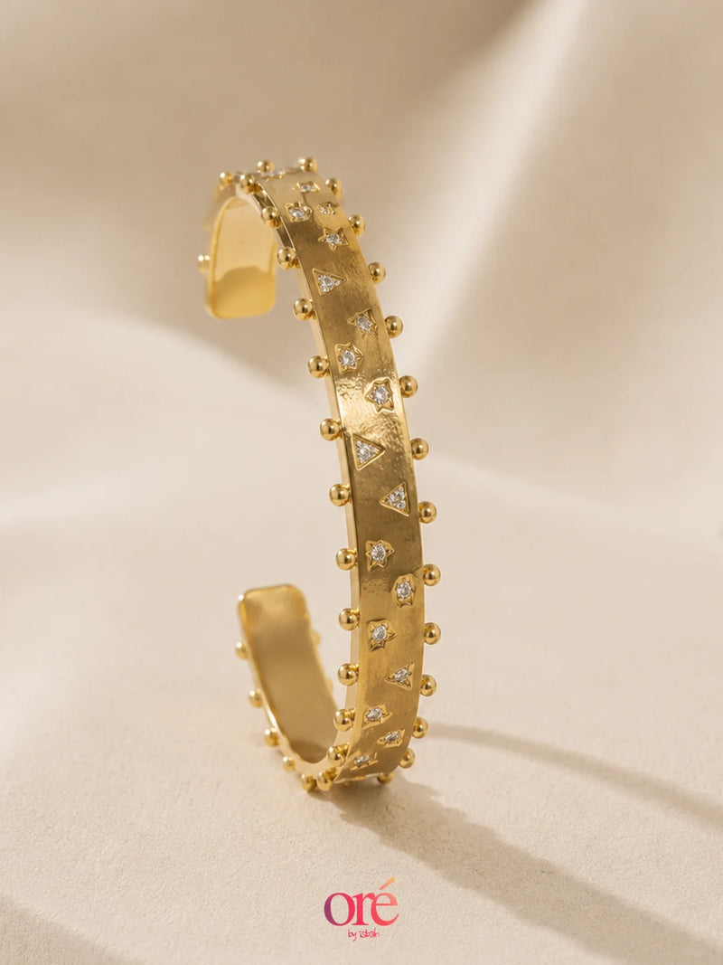 A gold-toned cuff bracelet with small clear stones and raised bead details, photographed on a soft beige fabric background. The brand name 'ore by ore' appears in pink at the bottom.