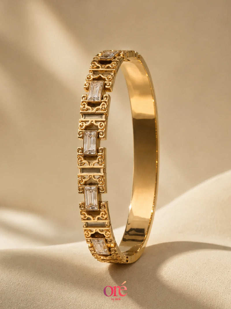 A gold-toned bangle bracelet with rectangular clear baguette and round-cut stones, displayed on a beige fabric surface with soft shadows. The brand name 'Ote Jewelry' is visible at the bottom.