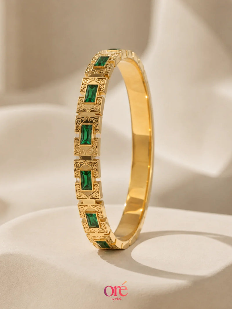 A gold bangle bracelet with rectangular green emerald-cut stones, displayed on a draped beige fabric background. The brand logo 'ore by ore' is visible in the bottom right corner.
