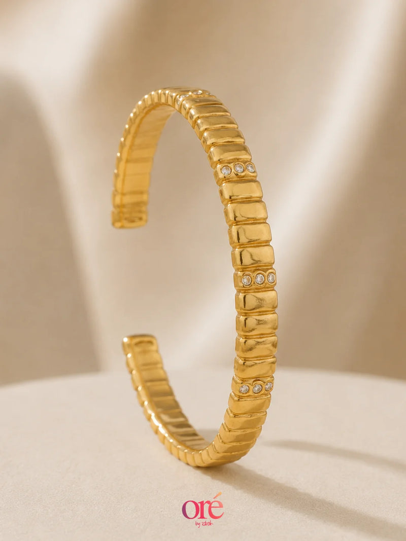 A gold-toned textured bangle bracelet with small clear rhinestone accents, displayed on a soft beige surface with a draped fabric background.