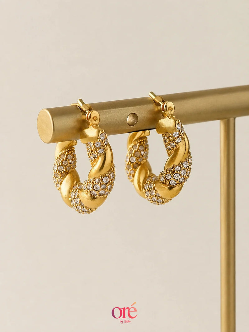 Gold hoop earrings with pavé-set clear stones, hanging from a gold display stand against a light beige background. A small 'ore by ore' logo watermark is visible at the bottom.