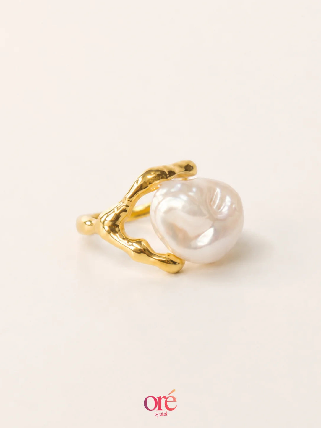 A gold ring featuring a large, irregular white baroque pearl nestled in a textured, organic-shaped gold setting, photographed on a clean off-white background with a brand watermark at the bottom.