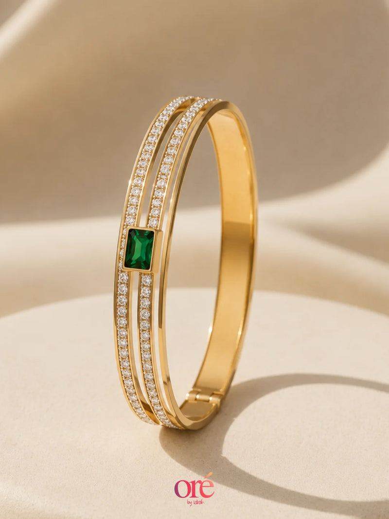 A gold-plated double-band bangle bracelet with a central square-cut green emerald accent and rows of small clear pavé stones, displayed on a beige fabric background.