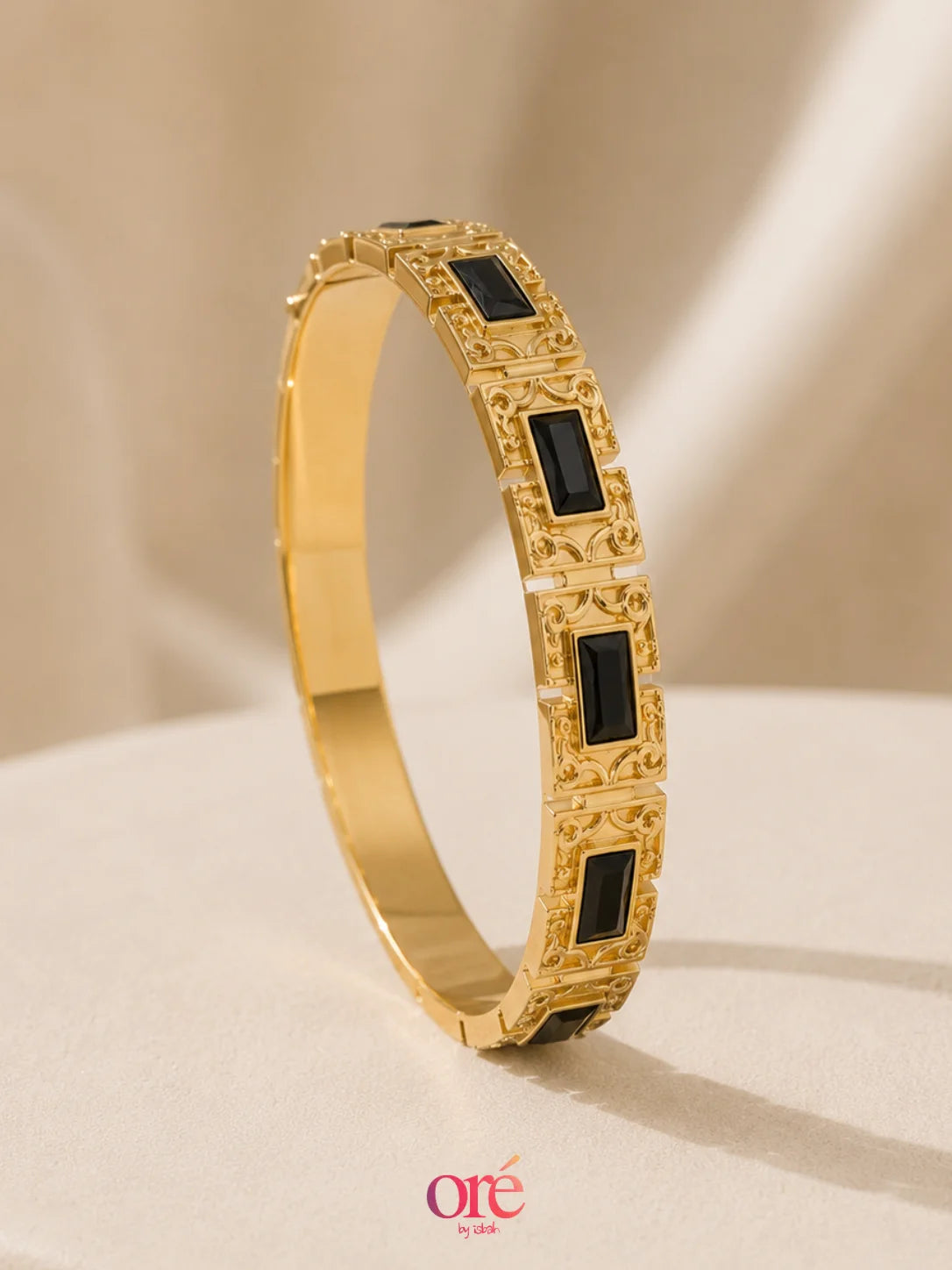 A gold hinged bangle bracelet with rectangular black inlay stones, displayed on a beige surface with a soft beige background. The brand logo 'ore by ore' is visible at the bottom.