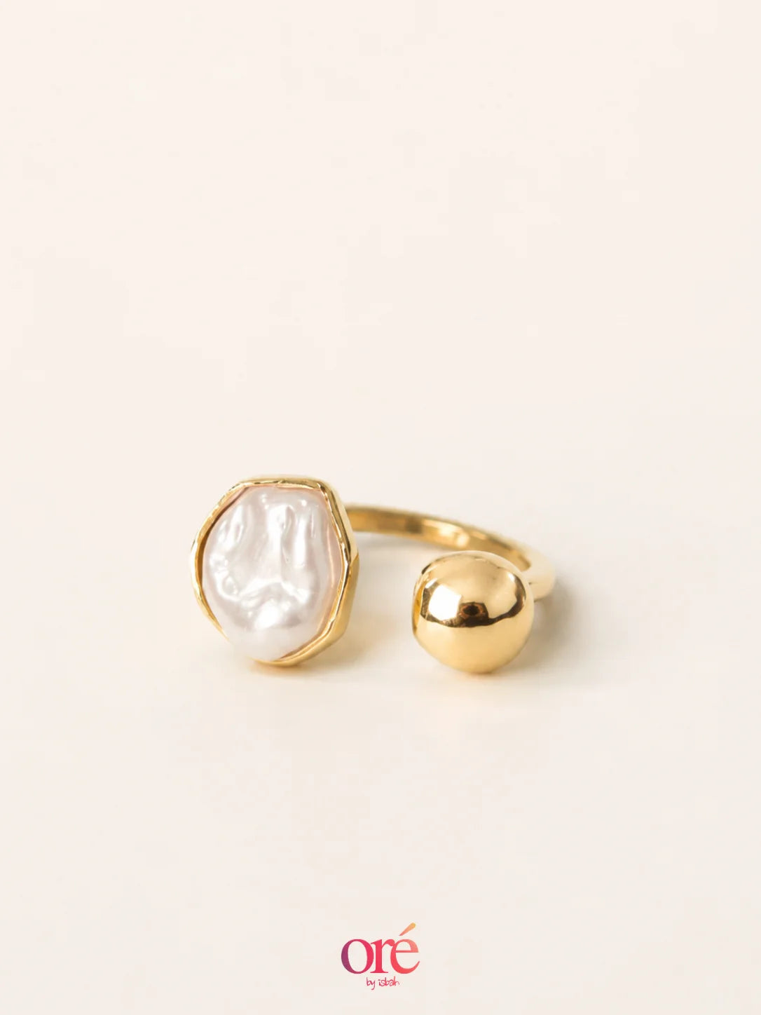 A gold ring featuring a large, irregularly shaped white pearl and a separate polished gold bead, set against a warm off-white background with an 'ore by ore' logo at the bottom.