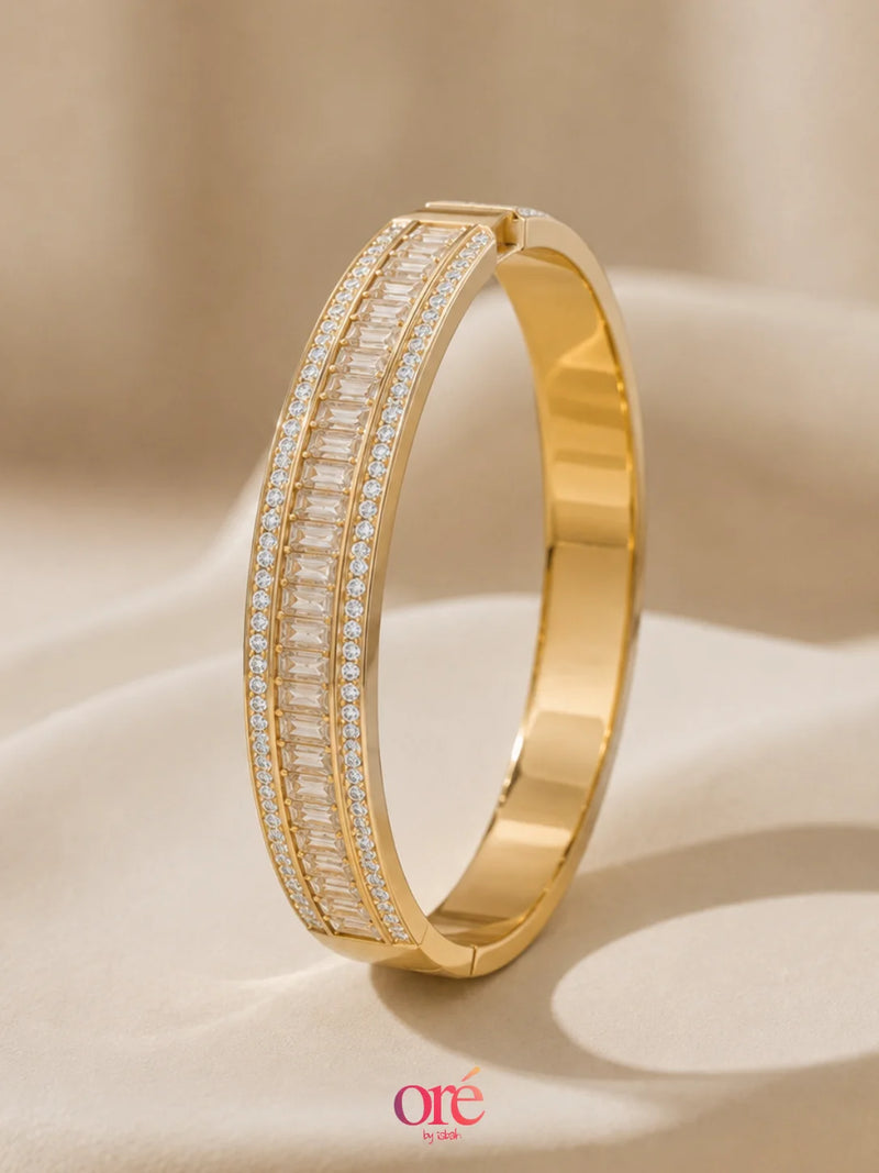 A polished gold hinged bangle bracelet featuring two parallel channels of baguette-cut clear stones, displayed on a draped beige fabric background.