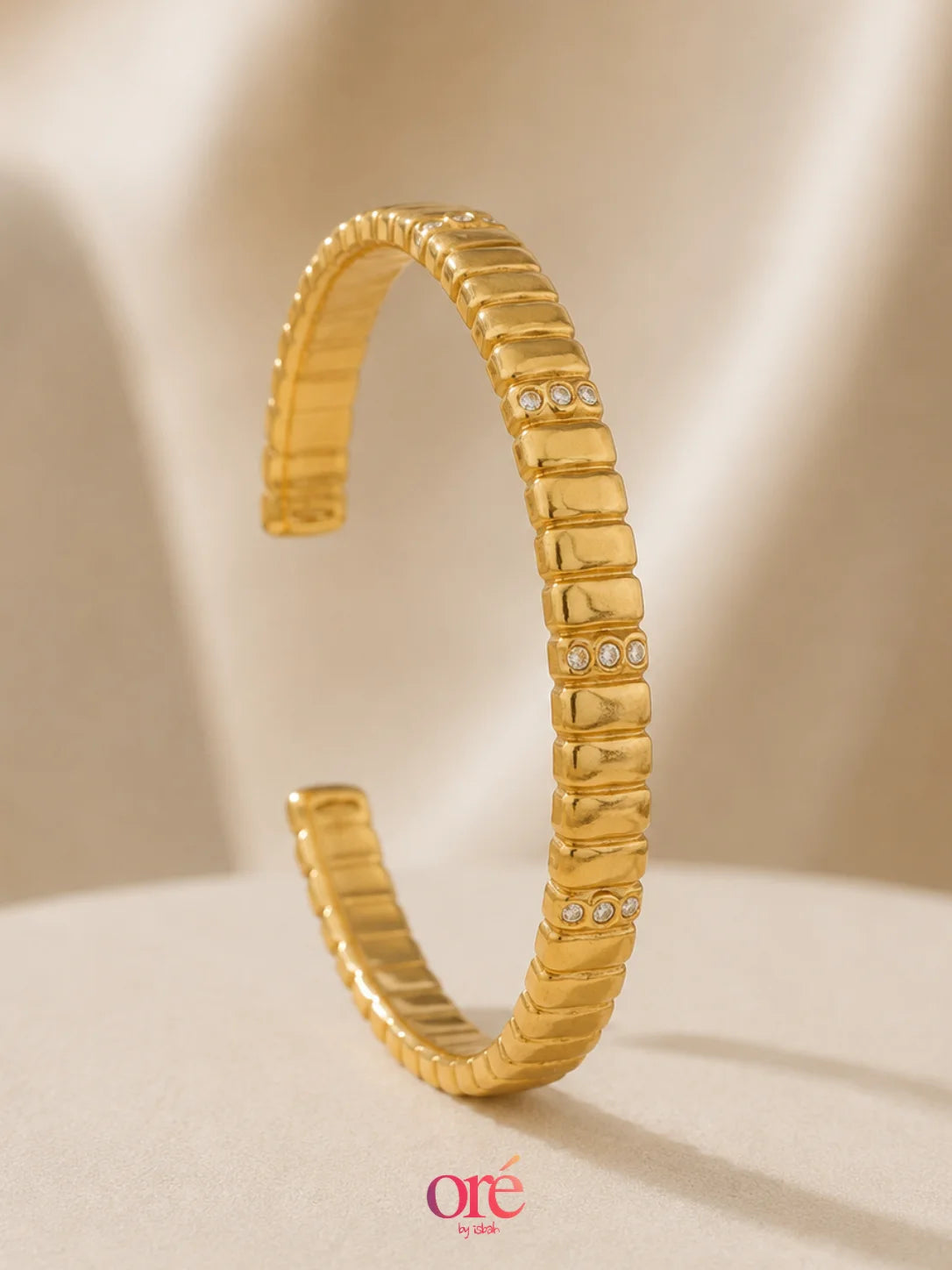 A gold-toned textured bangle bracelet with small clear rhinestone accents, displayed on a soft beige surface with a draped fabric background.