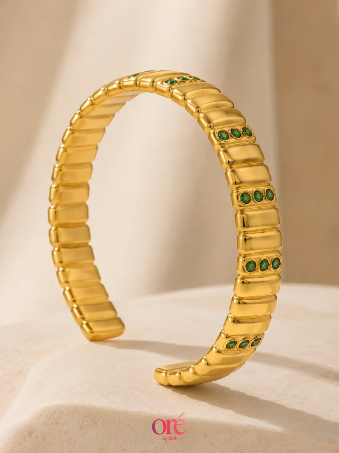A gold segmented cuff bracelet with small green stone accents, displayed on a beige fabric surface with a soft shadow.