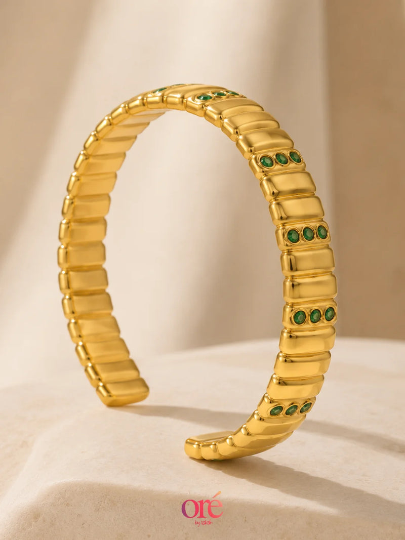 A gold segmented cuff bracelet with small green stone accents, displayed on a beige fabric surface with a soft shadow.