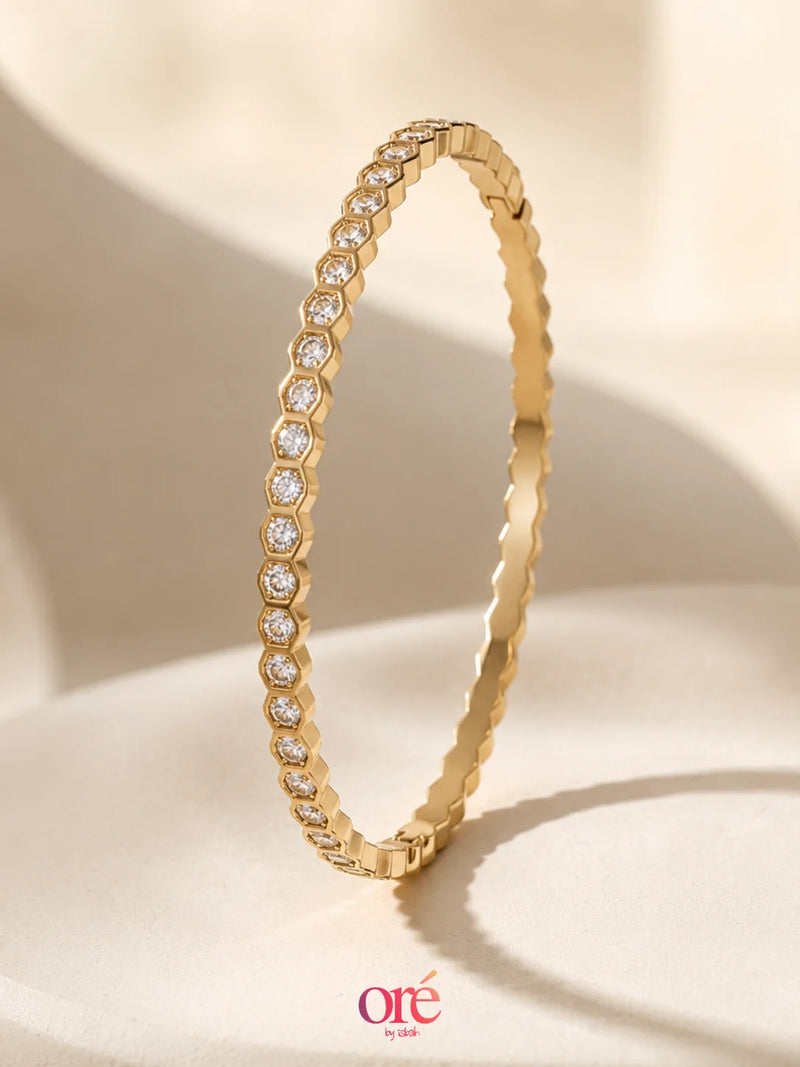A gold bangle bracelet with a row of clear pavé-set round stones, displayed on a soft beige fabric background. The brand logo 'ore by ore' is visible in the bottom right corner.