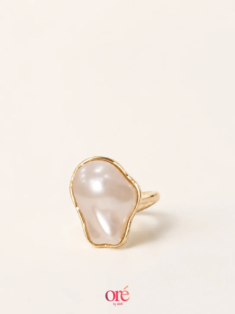 A gold ring featuring an organic, irregularly shaped white pearl, photographed on a clean, solid off-white background with a brand watermark at the bottom.