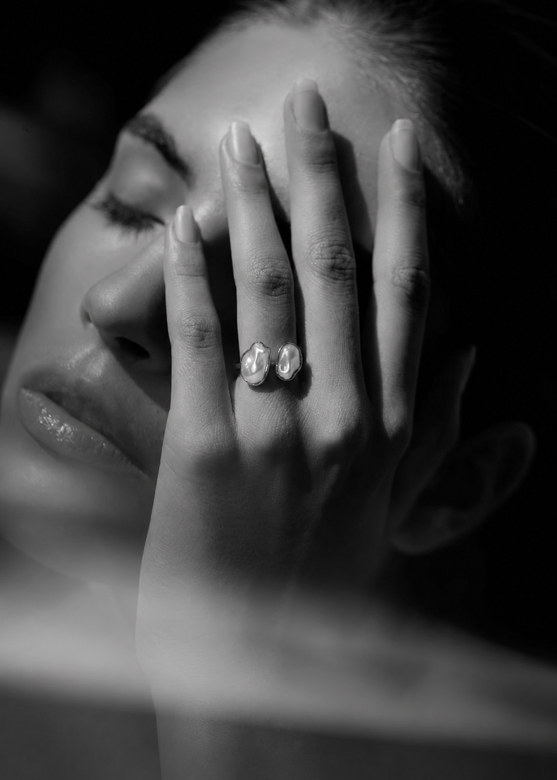 A close-up black and white photograph of a woman's face and hand, with two small oval stud earrings visible on her fingers.