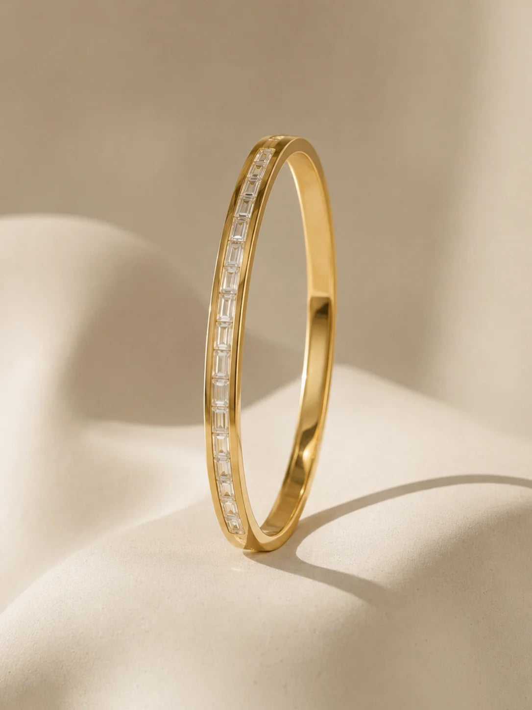 A polished gold hinged bangle bracelet featuring two parallel channels of baguette-cut clear stones, displayed on a draped beige fabric background.