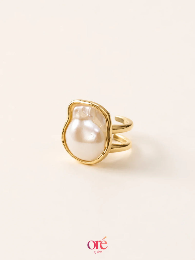 A gold ring featuring a large, irregular-shaped white pearl bezel-set in a polished gold frame, shown on a warm off-white background with an 'ore by ore' logo at the bottom.