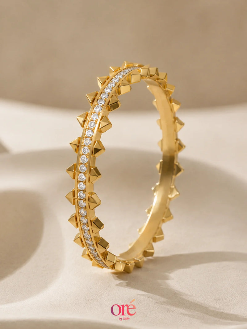 A gold-toned bangle bracelet with a raised spiked star pattern and a central row of clear stones, photographed on a soft beige surface. The brand name 'ore by ore' appears at the bottom.