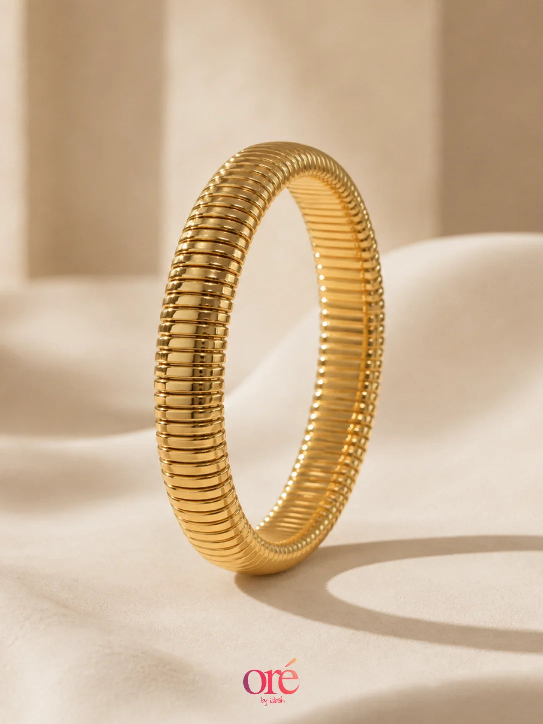 A gold snake-braided bangle bracelet standing upright on a draped beige fabric surface with a soft, warm background. The brand logo 'ore by ore' is visible in the bottom right corner.