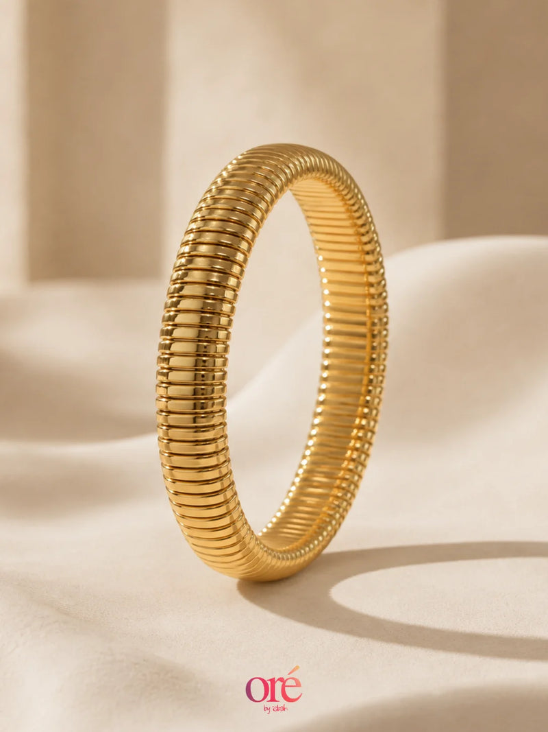 A gold snake-braided bangle bracelet standing upright on a draped beige fabric surface with a soft, warm background. The brand logo 'ore by ore' is visible in the bottom right corner.