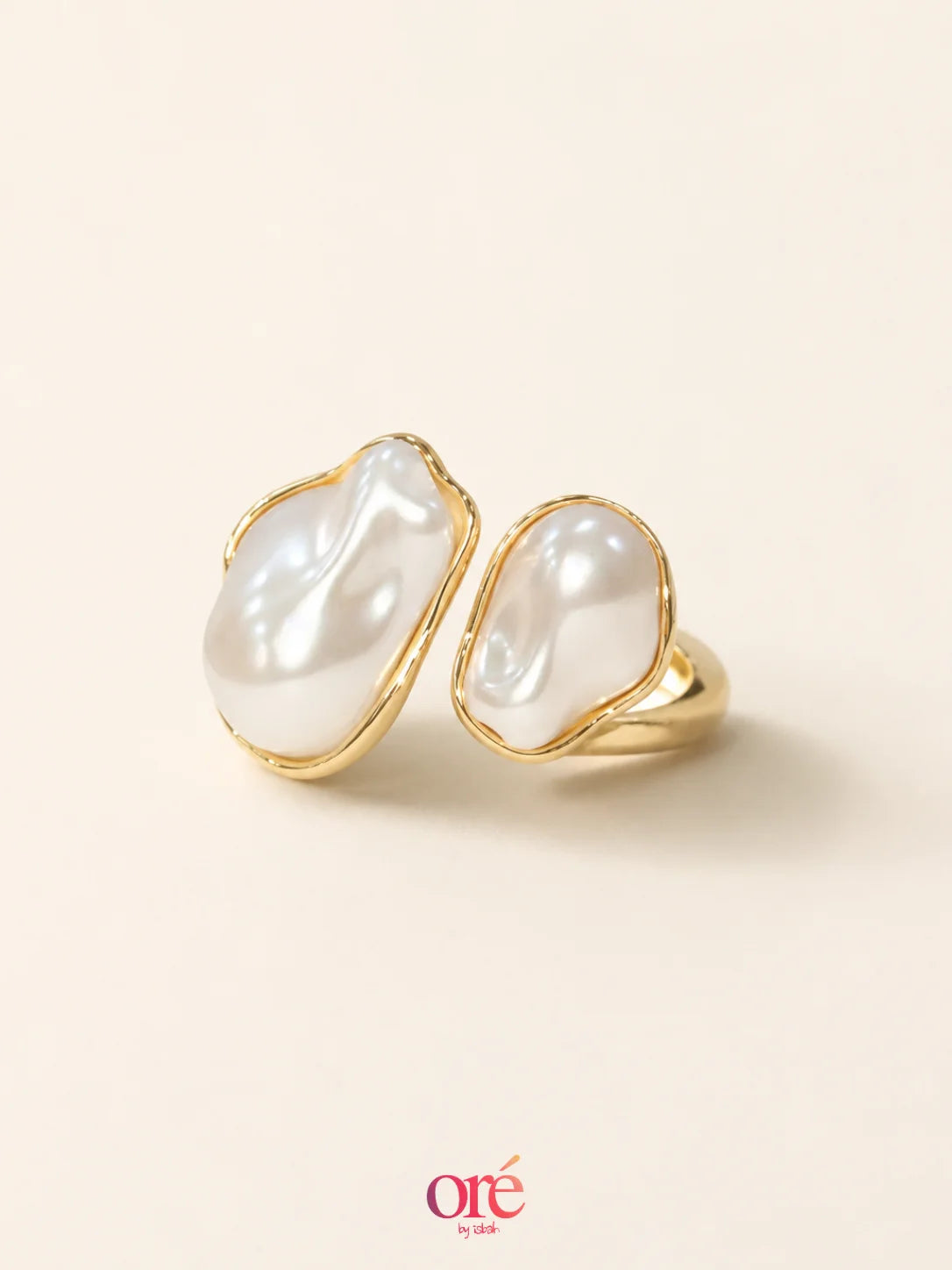 A pair of gold rings featuring large, organic-shaped white baroque pearls, displayed on a warm cream background with the 'ore by ore' logo at the bottom.