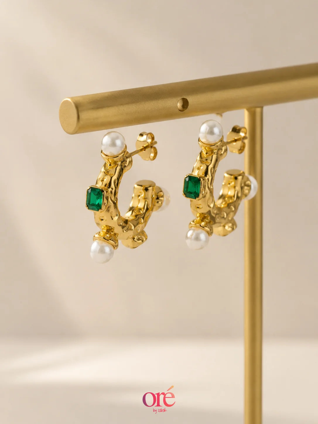 A pair of gold hoop earrings with white pearl accents and green rectangular gemstones, hanging from a gold display stand against a warm beige background.