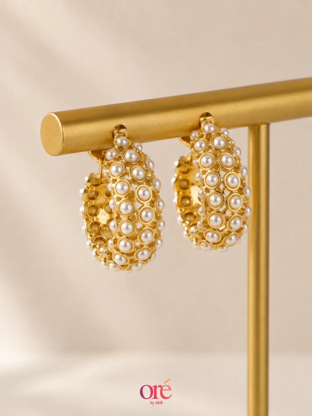 Gold hoop earrings adorned with small white pearls, displayed on a gold stand against a soft beige background. The brand logo 'ore by ore' is visible in the bottom corner.