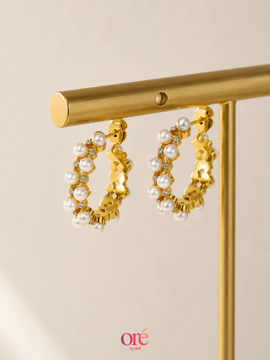 Gold hoop earrings adorned with white faux pearls and small clear rhinestones, hanging from a gold display stand against a warm beige background.