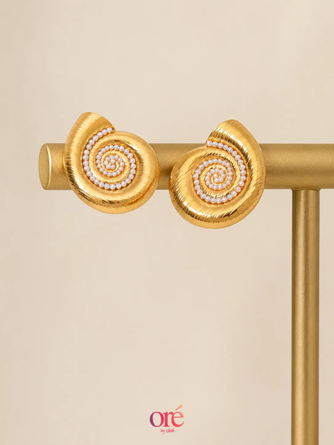 A pair of gold spiral stud earrings with a row of small white stones in the center, displayed on a gold stand against a warm beige background.