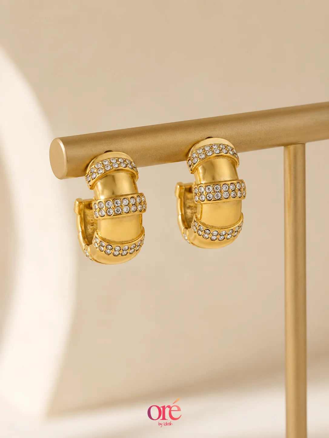 Gold hoop earrings with rows of clear pavé-set stones, displayed on a gold jewelry stand against a warm cream background. The brand logo 'ore by ore' is visible in the bottom right corner.