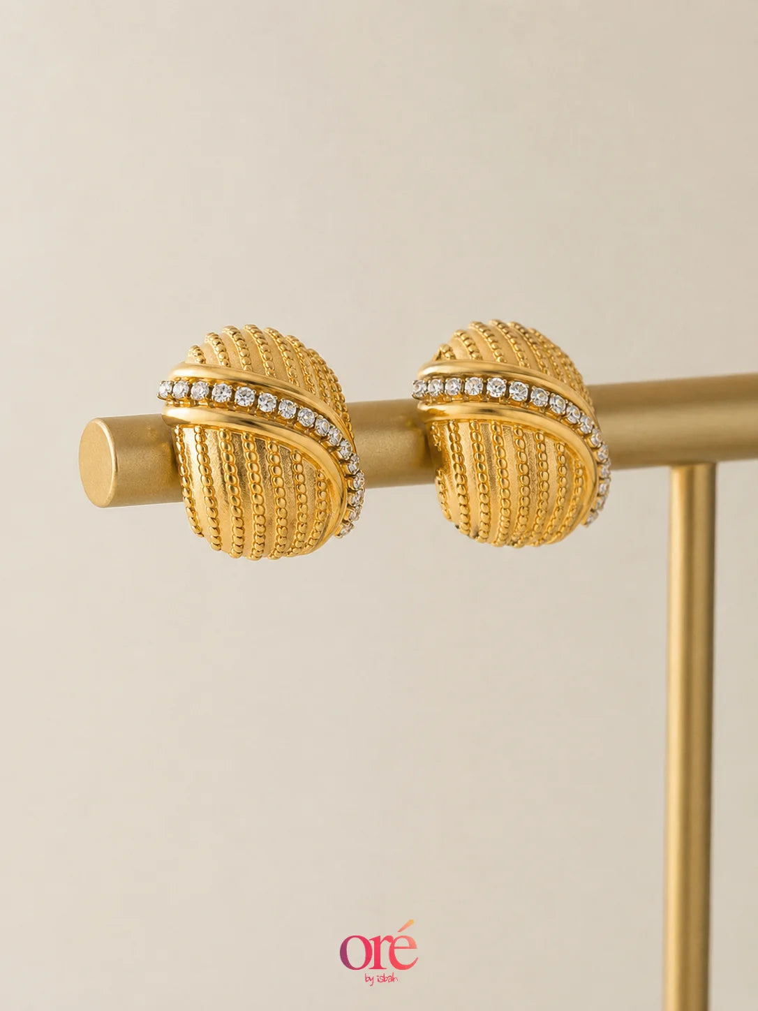 A pair of gold textured wrap earrings with a row of small clear stones, displayed on a gold stand against a light beige background.