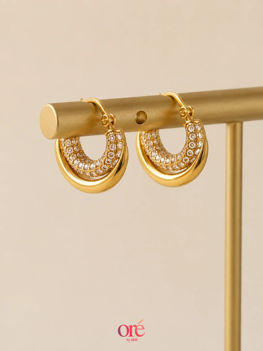 Gold crescent-shaped hoop earrings with pavé-set clear stones, displayed on a gold jewelry stand against a warm beige background. The brand logo 'ore by ore' is visible at the bottom.